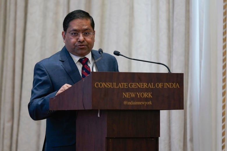 A man in a blue suit with black hair and black glasses speaks at a podium.