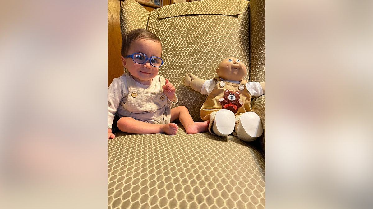 Baby Liam sits next to a doll on an armchair.