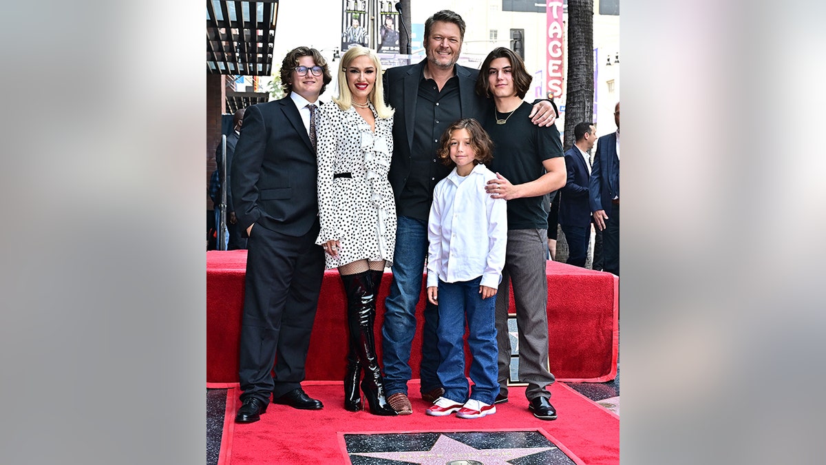 Gwen Stefani, in a two piece set, stands on the carpet with Blake Shelton in a black suit and blue jeans and her three sons at Shelton's Walk of Fame ceremony